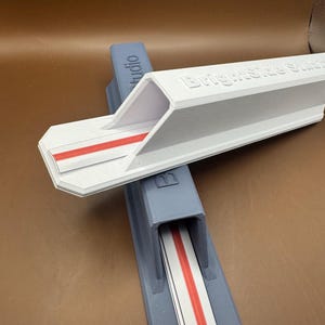 May include: Two 3D-printed plastic track components, one white and one grey, each featuring a central red stripe. The white component is embossed with the text Brightside Studio. Both pieces have a hollow, angled, elongated rectangular design.