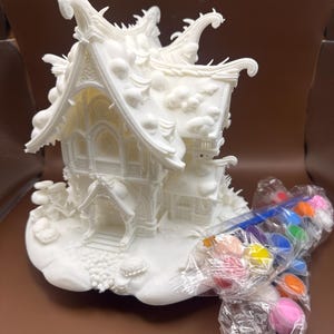 May include: A white, detailed miniature house with an ornate roof and architectural details. A set of colorful paint pods and a brush are included. The house is designed for painting and crafting.