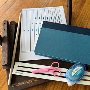 May include: A wooden bookbinding kit with a blue notebook, a pink pair of scissors, a white tape dispenser, and a white sheet of paper with a grid pattern. The kit is labeled "Crafted Sweetly".
