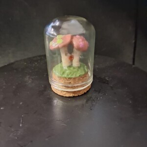 May include: A miniature mushroom diorama inside a clear glass cloche. The mushrooms are pink with green and white accents, set on a green base. The base is made of cork. The cloche is on a dark surface.