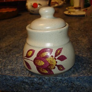 Peut inclure: Un pot à sucre en céramique blanche avec un couvercle, orné d'un motif floral peint à la main dans des tons de rose, jaune et vert. Le pot a un glaçage moucheté et une forme arrondie.