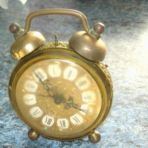 May include: An antique brass alarm clock with a round face and Roman numerals. The clock has a handle and two bells on top. The clock's face is a light gold color, and the hands are black. The clock is resting on a blue surface.
