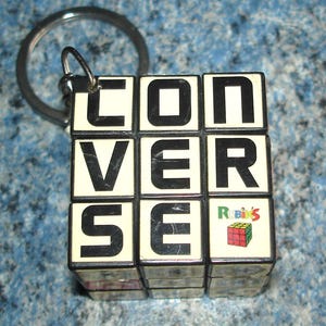 Peut inclure: Un porte-clés noir et blanc en forme de Rubik's Cube avec le mot "CONVERSE" écrit sur le devant. Le porte-clés est doté d'un anneau de clé argenté. Le porte-clés Rubik's Cube porte le logo "Rubik's" sur le côté.
