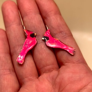 Op de afbeelding: Een paar roze kardinaal oorbellen met zilveren hoepels. De kardinalen zijn geschilderd met een zwarte omtrek en hebben een glanzende afwerking.