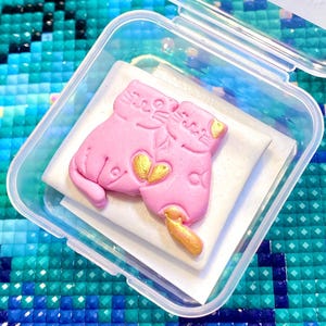 Puede incluir: Un dije rosa con forma de gato con detalles dorados, colocado sobre un cuadrado blanco dentro de un recipiente de plástico transparente. El dije presenta dos gatos con un diseño en forma de corazón. El fondo es un mosaico de cuadrados azules y verdes.