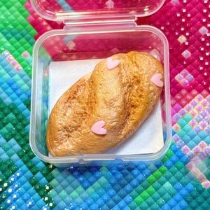 Puede incluir: Una barra de pan con decoraciones rosas en forma de corazón dentro de un recipiente de plástico transparente. El pan tiene una corteza dorada y marrón y está sobre papel blanco. El recipiente está abierto y el fondo es un patrón de mosaico colorido.