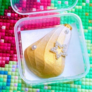 Puede incluir: Un objeto decorativo dorado y blanco con forma de concha, adornado con una estrella de perlas y perlas individuales. El artículo está dentro de un recipiente de plástico transparente con un forro de papel blanco. El fondo es un mosaico colorido.