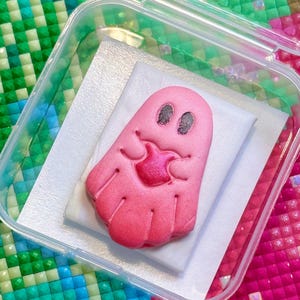 May include: A small, pink, ghost-shaped figurine with dark, textured oval eyes and a central, heart-shaped mouth detail. The figurine is placed on a white rectangular backing inside a clear, square plastic storage container with a hinged lid.