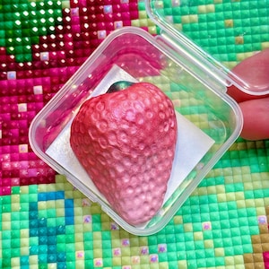 Puede incluir: Una escultura realista de fresa roja con superficie texturizada, exhibida en un recipiente de plástico transparente. La fresa tiene un degradado de color, de rojo a rosa. El recipiente está sobre un cuadrado blanco.