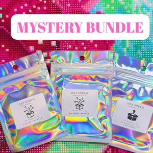 Puede incluir: Tres bolsas iridiscentes con la etiqueta "Mystery Bundle", el texto "The Classic Line" y "THES STUDIOS". El fondo es un colorido patrón de mosaico. Cada bolsa tiene una pequeña etiqueta blanca con una caja y signos de interrogación.