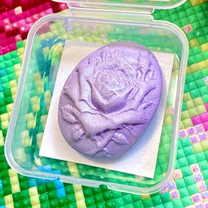 Puede incluir: Una bomba de baño ovalada de color lavanda con un diseño de rosa, sobre papel blanco dentro de un recipiente de plástico transparente. La bomba de baño tiene una superficie texturizada y un brillo sutil. El recipiente está abierto.