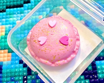 May include: A pink, heart-shaped dessert with three smaller pink hearts on top, inside a clear plastic container. The dessert has gold flecks and a ruffled edge. The container sits on a white paper square.