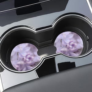 May include: Two circular car cup holder inserts featuring a soft-focus, light purple floral pattern. The coasters are placed inside a black plastic dual cup holder console with a glossy finish, designed to fit standard vehicle interior spaces.