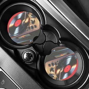 May include: Black car cup holders with two round coasters featuring a vintage record player design. The coasters have a red and black vinyl record on a turntable, with a retro aesthetic.