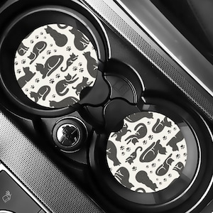 May include: Two car cup holder coasters with a black and white cat pattern. The coasters feature various cat silhouettes and paw prints on a cream background. The coasters are round and fit inside a car's cup holders.