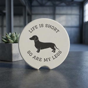 May include: A cream-colored car coaster with a black dachshund silhouette and the text "LIFE IS SHORT SO ARE MY LEGS" in a circular design. The coaster has a small notch on one side.