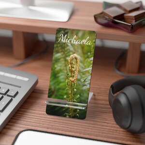 Puede incluir: Un soporte para teléfono con un diseño temático de la naturaleza. El soporte presenta una imagen vibrante de un helecho con el nombre "Michaela" en una escritura elegante. El soporte está hecho de acrílico transparente y está diseñado para sostener un teléfono o una tableta pequeña.