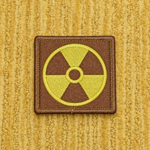 May include: A square embroidered patch featuring a brown background with a yellow radiation symbol centered in the middle. The patch has a stitched border in a matching brown thread, creating a clean edge around the square shape of the design.