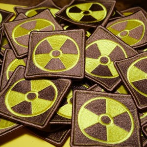 Może przedstawiać: Stos brązowych i żółtych kwadratowych łat z wyszytym na każdej z nich żółtym symbolem promieniowania.