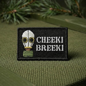 Może przedstawiać: Czarna, prostokątna, haftowana naszywka z motywem maski gazowej w kolorze białym, żółtym i zielonym. Tekst "CHEEKI BREEKI" jest wyhaftowany na biało. Naszywka jest przeznaczona do mocowania na odzieży lub akcesoriach.