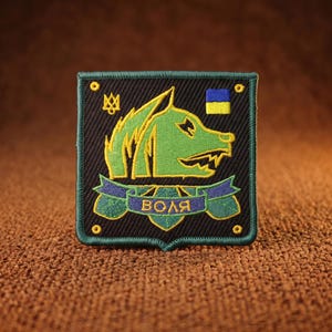 May include: A square embroidered patch with a black background and a green wolf head silhouette. It features a small yellow Ukrainian trident symbol, a blue and yellow flag, and a blue ribbon banner with the Cyrillic text "ВОЛЯ" in yellow.