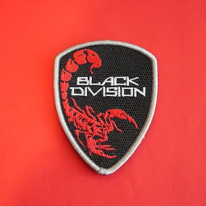 May include: A black and red embroidered patch with the words "BLACK DIVISION" and a red scorpion design. The patch is shield-shaped with a silver border, set against a red background.