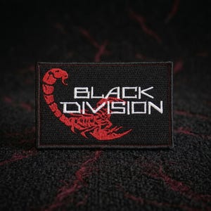 May include: A rectangular black embroidered patch featuring a red scorpion graphic on the left side and the white text "BLACK DIVISION" on the right side. The patch has a thin red border and a textured fabric finish with visible stitching.
