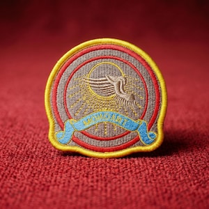 May include: Embroidered patch featuring a stylized bird with outstretched wings, a sunburst design, and the word "MOHOAMIT" on a blue banner. The patch has a gray background with red and yellow borders, set against a red fabric.