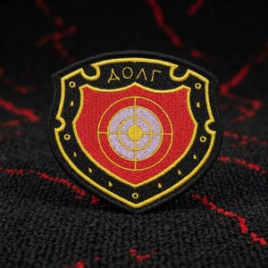 May include: Embroidered patch in a shield shape with a red background and a target design in the center. The patch has a black border with gold accents and the word "ДОЛГ" in gold.