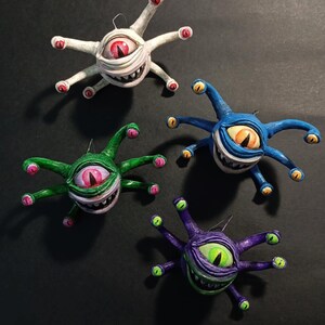 Può includere: Quattro ornamenti fatti a mano che raffigurano una creatura con più occhi e un ghigno pieno di denti. Gli ornamenti sono dipinti in diversi colori: bianco, verde, blu e viola.