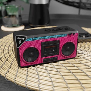 May include: A rectangular Bluetooth speaker designed to resemble a retro boombox. The device features a bright pink perforated speaker grille, a central black cassette tape graphic, a teal accent strip, and a black Bluetooth logo on the side.