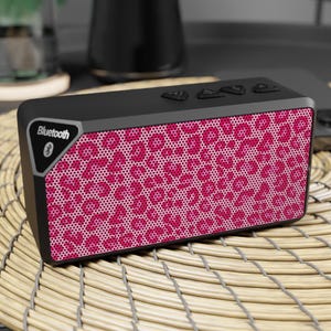 May include: A rectangular Bluetooth speaker featuring a vibrant pink and black leopard print pattern on the front grille. The device has a black frame with a triangular accent on the top left corner displaying the word Bluetooth and a logo.