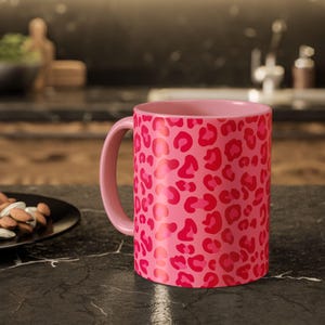 May include: A ceramic mug featuring a vibrant pink and red leopard print pattern. The mug has a smooth, cylindrical body with a matching pink handle on the left side. The design covers the entire exterior surface of the mug in a repeating animal print.