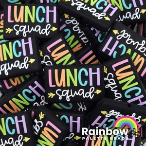 May include: A close-up of a pile of black silicone charms with the text "Lunch Squad" in various colors. The charms are in a variety of shapes and sizes.