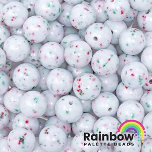 May include: A collection of white, round beads with red and green speckles. Each bead has a hole for stringing. The Rainbow Palette Beads logo, featuring a rainbow, is visible in the lower right corner.