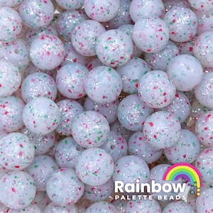 May include: A close-up of a pile of white silicone beads with red and green glitter. The beads are round and have a speckled appearance. The beads are arranged in a random pattern. The beads are for use in jewelry making or as sensory toys.