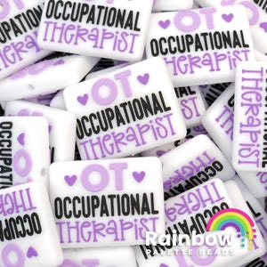 May include: A close-up of a pile of white, rectangular, silicone beads with the text "OT Occupational Therapist" in black and purple lettering. The beads have small purple hearts on them.