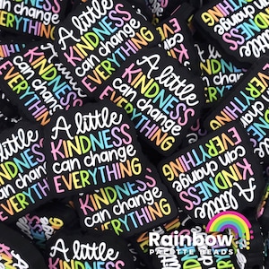 May include: A close-up of a pile of colorful silicone charms with the text "A little kindness can change everything" repeated on each charm. The charms are black with rainbow lettering.