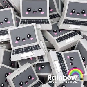 May include: A pile of gray laptop-shaped silicone charms with cute cartoon faces. Each charm has a black keyboard and a white screen. The charms are arranged in a random pattern. The charms are from Rainbow Palette Beads.