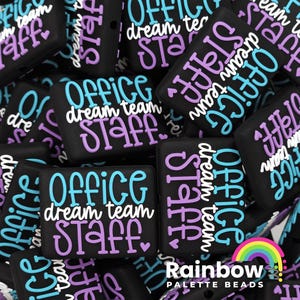 May include: A close-up of several black silicone square beads with the text "Office Dream Team Staff" in white, purple, and blue lettering. The beads are scattered on a surface.