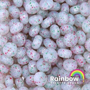 May include: A close-up of a large quantity of white donut beads with red and green glitter. The beads are scattered randomly and have a glossy finish.