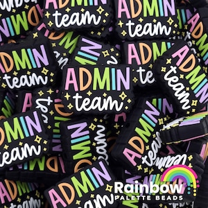 May include: A close-up of a pile of black silicone charms with the text "Admin Team" in rainbow colors. The charms are designed to be used on bracelets, necklaces, or keychains.