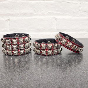 May include: Three black leather studded bracelets with silver pyramid studs and red accents. The bracelets have a punk rock aesthetic and are displayed on a dark gray surface.