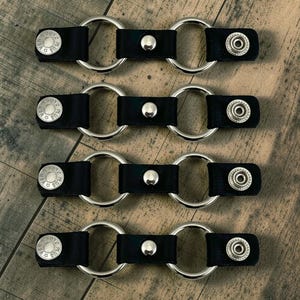May include: Four black leather straps, each featuring two silver-toned metal rings connected by a central snap button. Each end of the strap has a silver-toned snap fastener, with the left side embossed with the text USA PUNK PLUS.