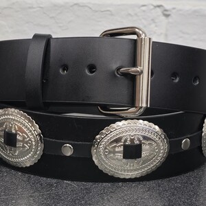 May include: A black leather belt with silver concho accents. The belt has a silver buckle and is decorated with four large, ornate silver conchos.