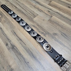 May include: A black leather belt with silver metal concho accents. The belt has a buckle with three prongs.