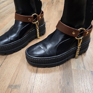 Puede incluir: Botas de cuero negro con suela gruesa y acanalada. Cada bota presenta una correa de tobillo de cuero marrón con herrajes dorados y una cadena dorada colgante. Sobre una superficie de madera marrón claro.