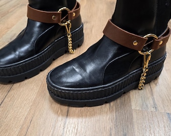 May include: Black leather boots with a thick, ridged sole. Each boot features a brown leather ankle strap with gold-colored hardware and a dangling gold chain. The boots are on a light brown wooden surface.
