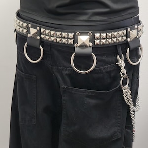 Könnte beinhalten: Schwarzer Ledergürtel mit silbernen Pyramiden-Nieten und großen silbernen Ringen. Der Gürtel hat eine Kette mit einem Verschluss. Der Gürtel wird mit einer schwarzen Hose getragen.