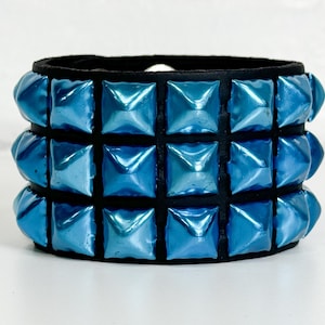 Puede incluir: Un brazalete de cuero negro con tres filas de tachuelas azules en forma de pirámide.